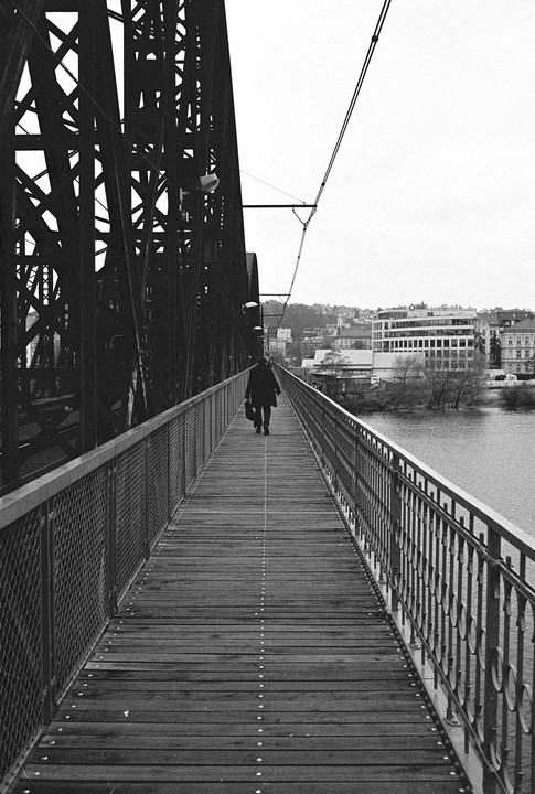 A person walks away from the viewer on a wooden bridge next to a river with buildings in the background.