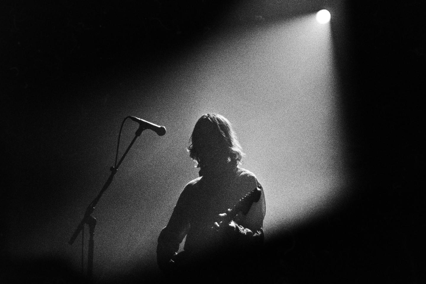 A musician with long hair plays a guitar on a stage under a spotlight next to a microphone.