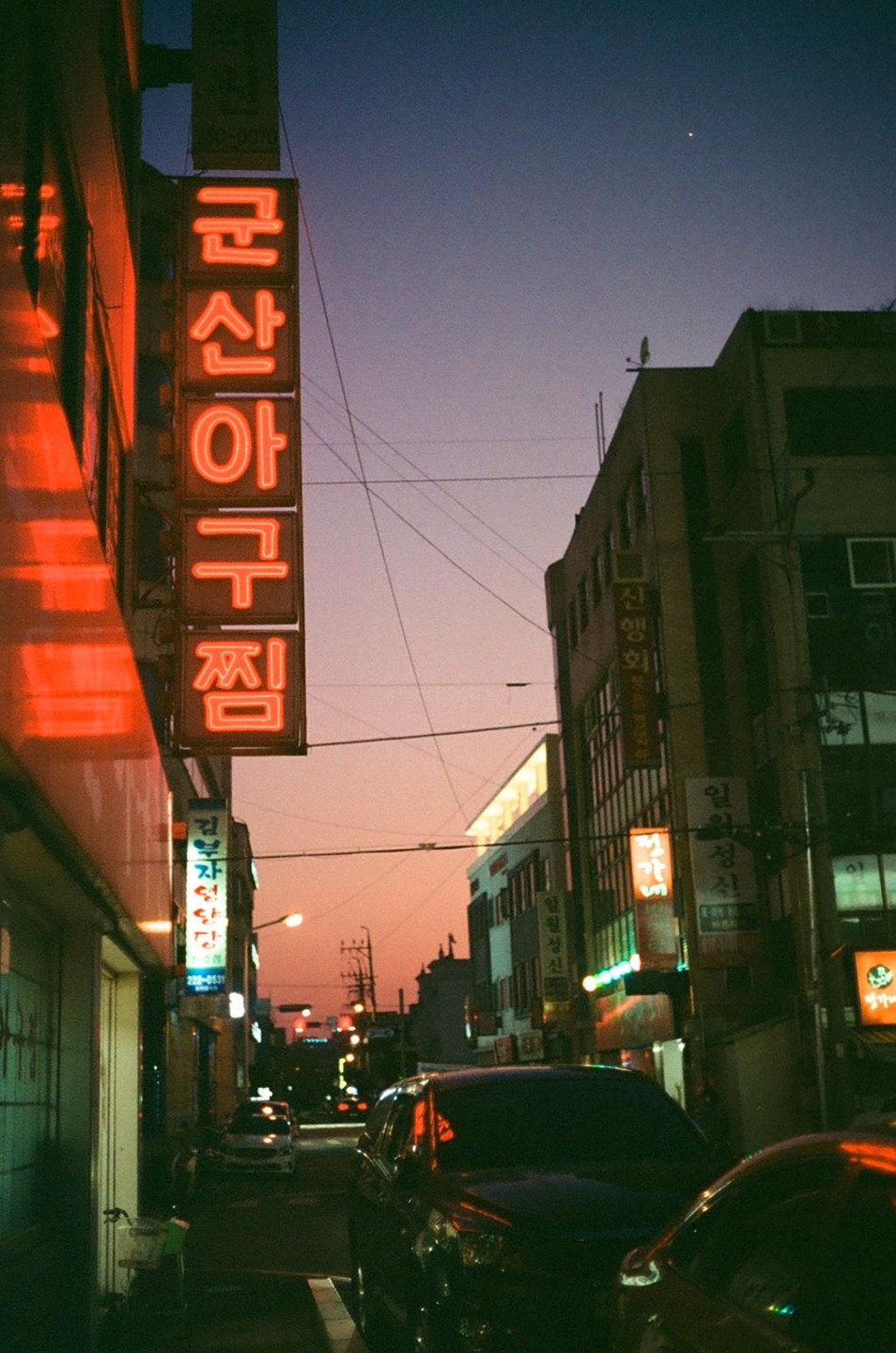 A street with neon signs and parked cars is visible at dusk.