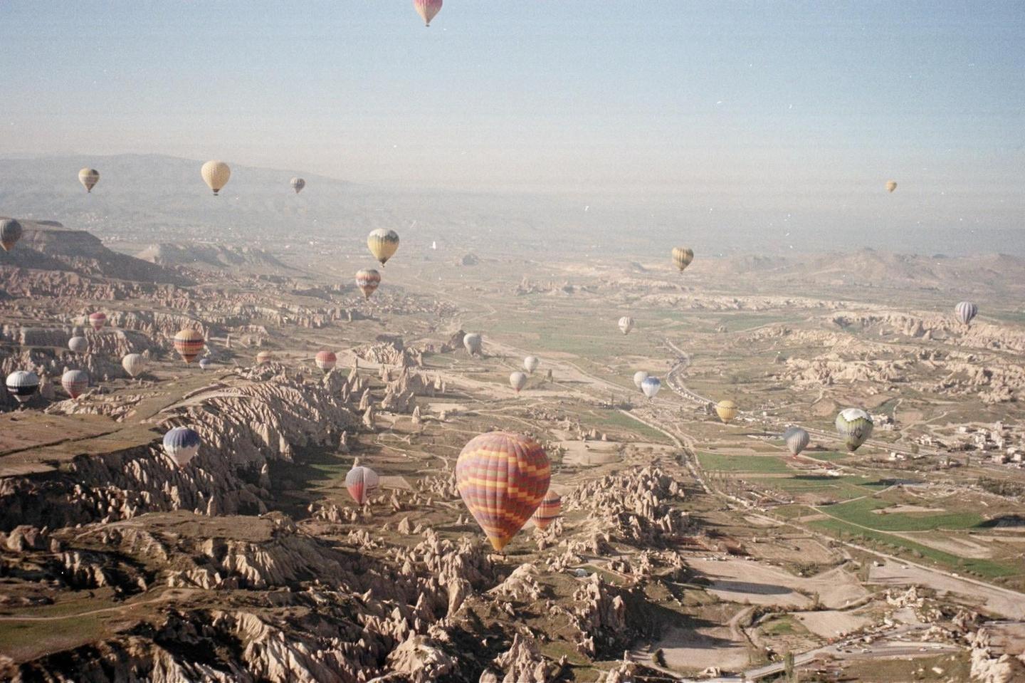 Hot air balloons float above a rugged, arid landscape with some green fields and a small town.