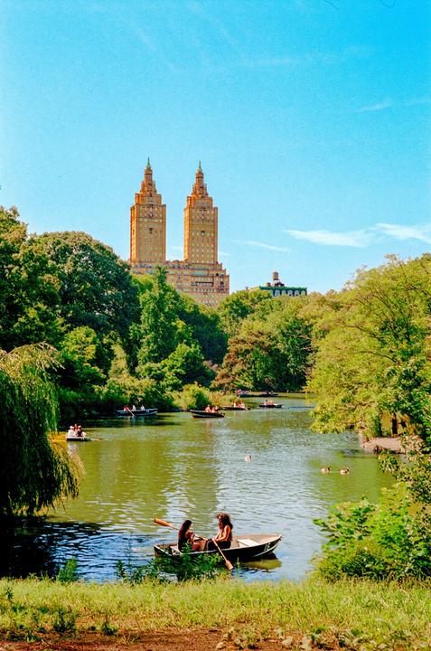 Two women row a boat on a lake with many other boats, surrounded by trees and buildings in the distance.