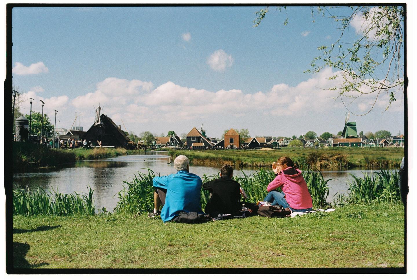 Three people sit on a grassy bank overlooking a canal with traditional Dutch buildings and a windmill in the background.