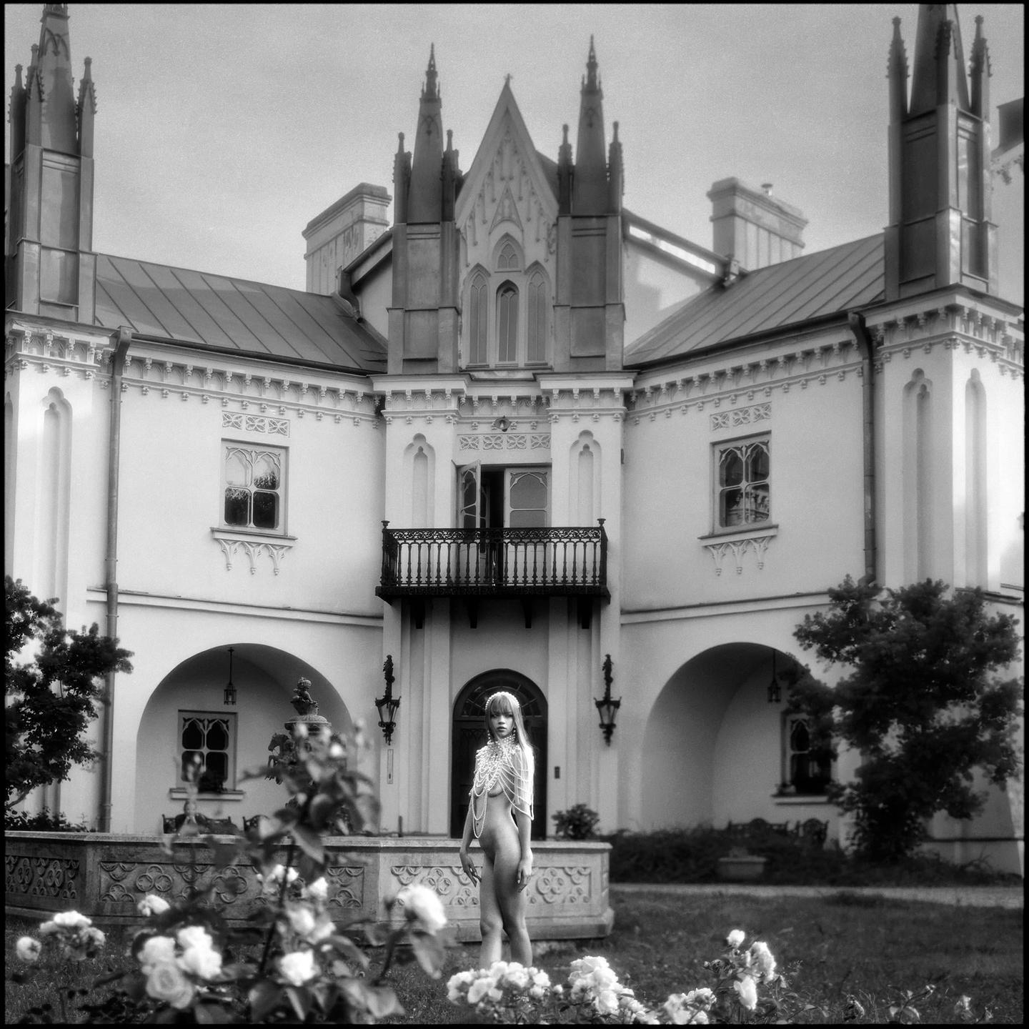 A nude woman wearing a pearl necklace stands in front of a gothic revival building with a garden in the foreground.