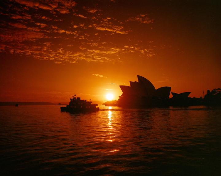 The Sydney Opera House and a boat are silhouetted against a vibrant red and orange sunset over the water.