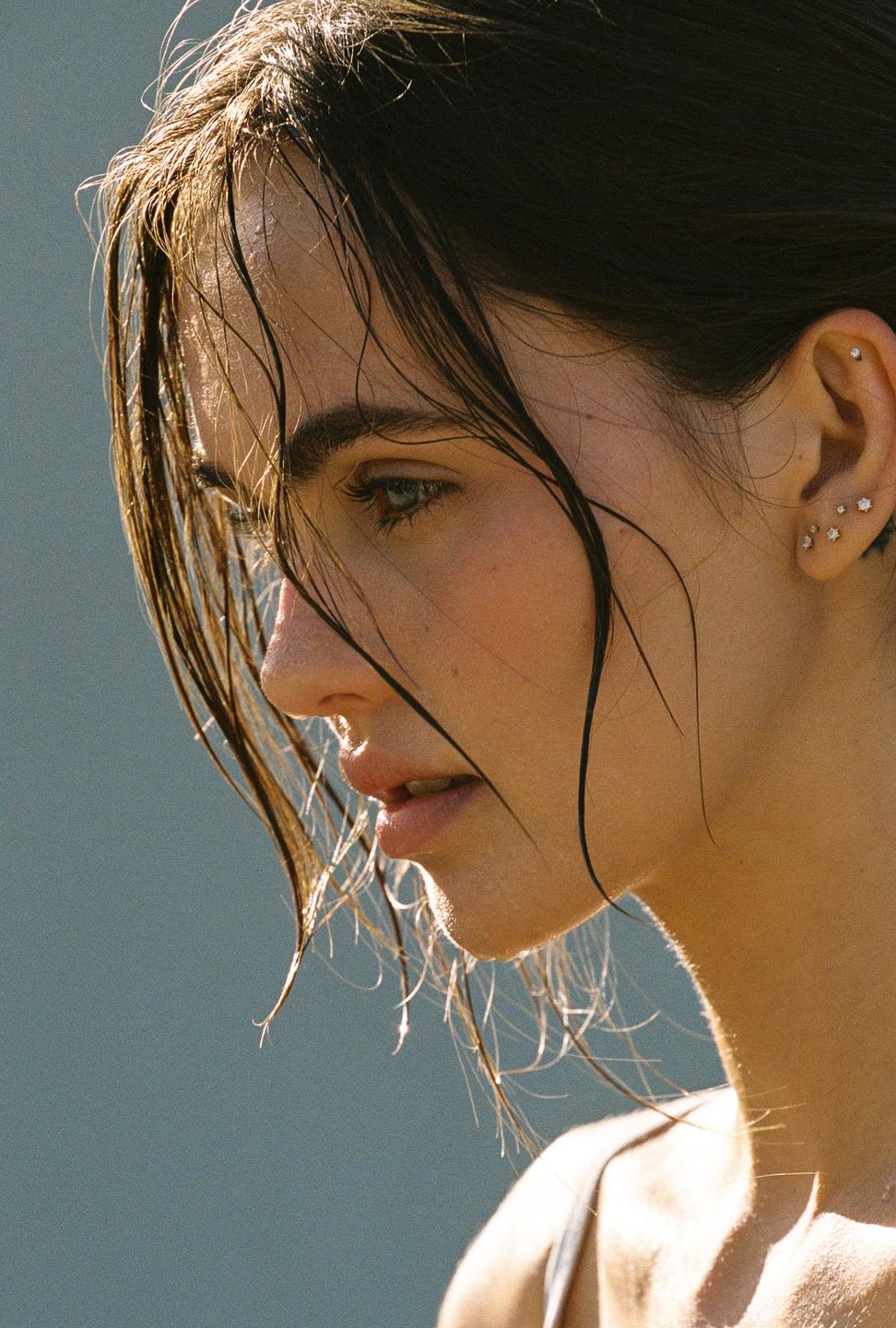 A woman with wet hair and multiple ear piercings looks down and to the left.