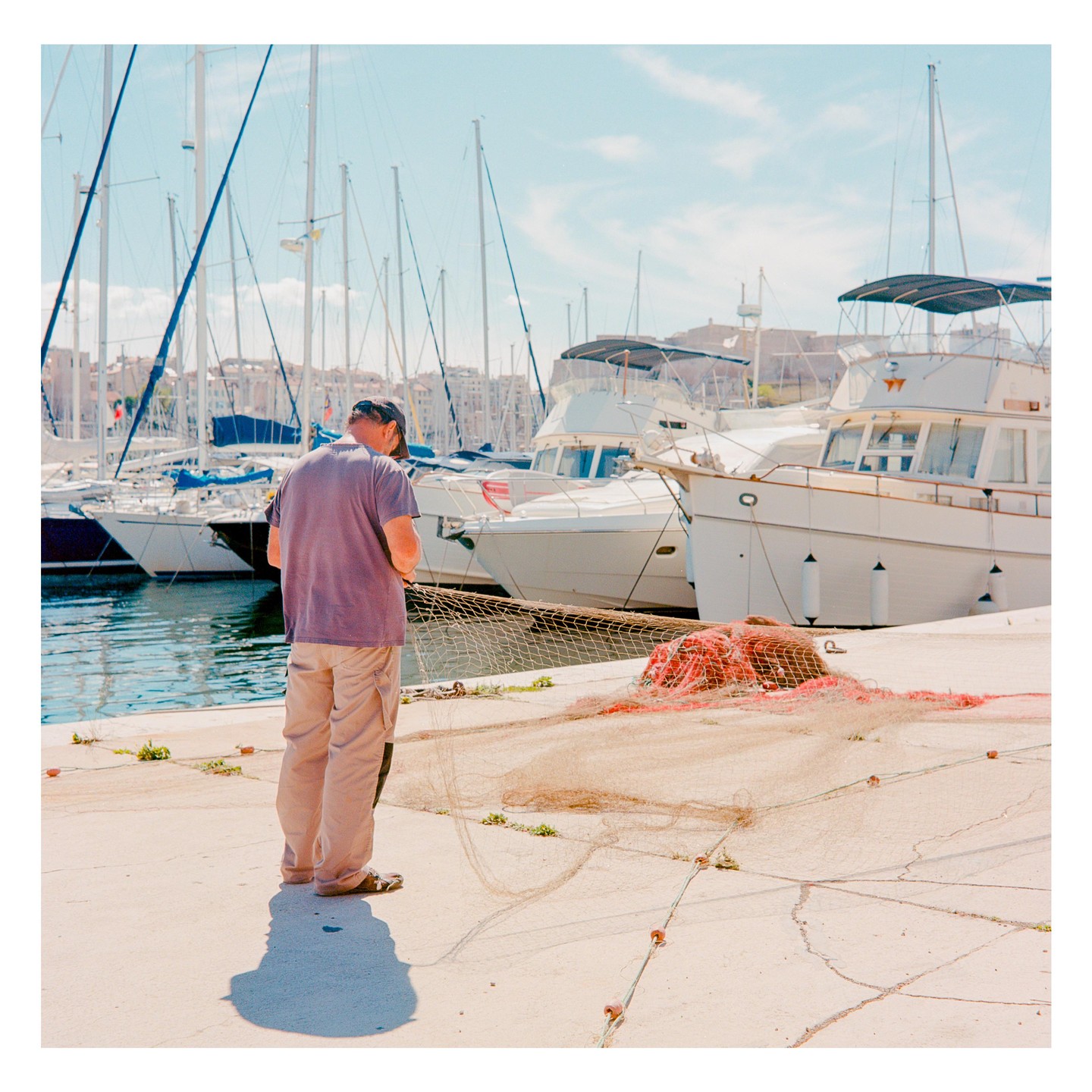 A man stands on a dock next to fishing nets with boats and buildings in the background.