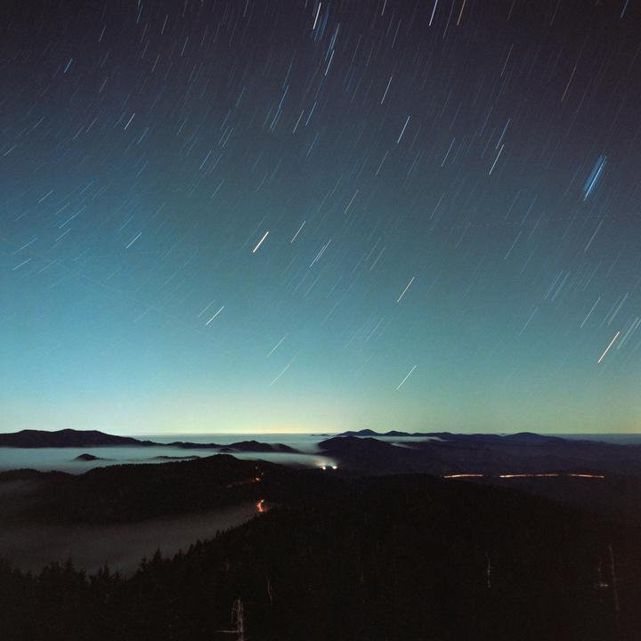 Star trails streak across the night sky above a mountain landscape with fog in the valleys.