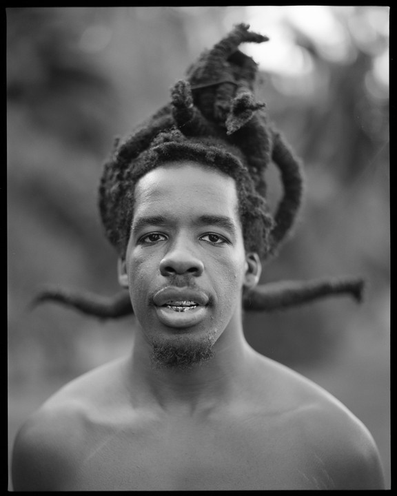 A man with dreadlocks and a goatee looks directly at the viewer.