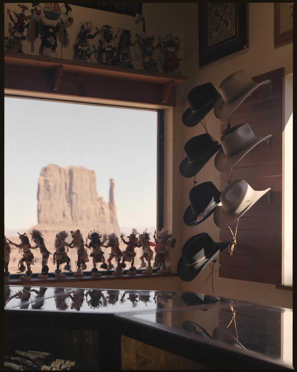 A window in a gift shop looks out onto a desert landscape with a large rock formation, and several cowboy hats hang on a wall to the right.