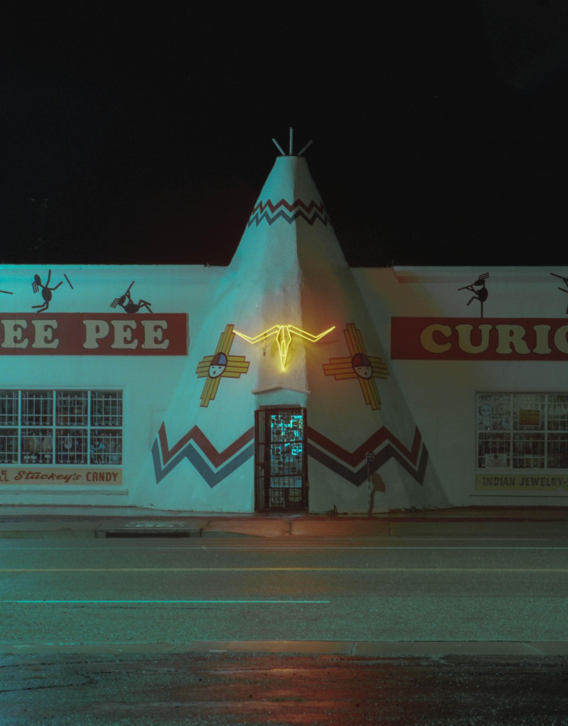 A teepee-shaped building with a neon bull skull sign stands beside a road at night.