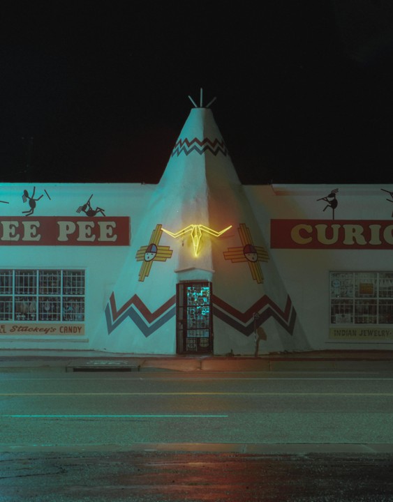 A teepee-shaped building with a neon bull skull sign stands beside a road at night.