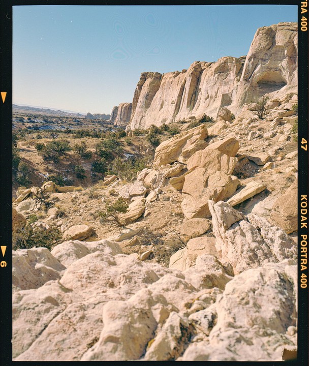 A vast desert landscape features a rocky foreground leading to a canyon with a cliff face under a clear sky.