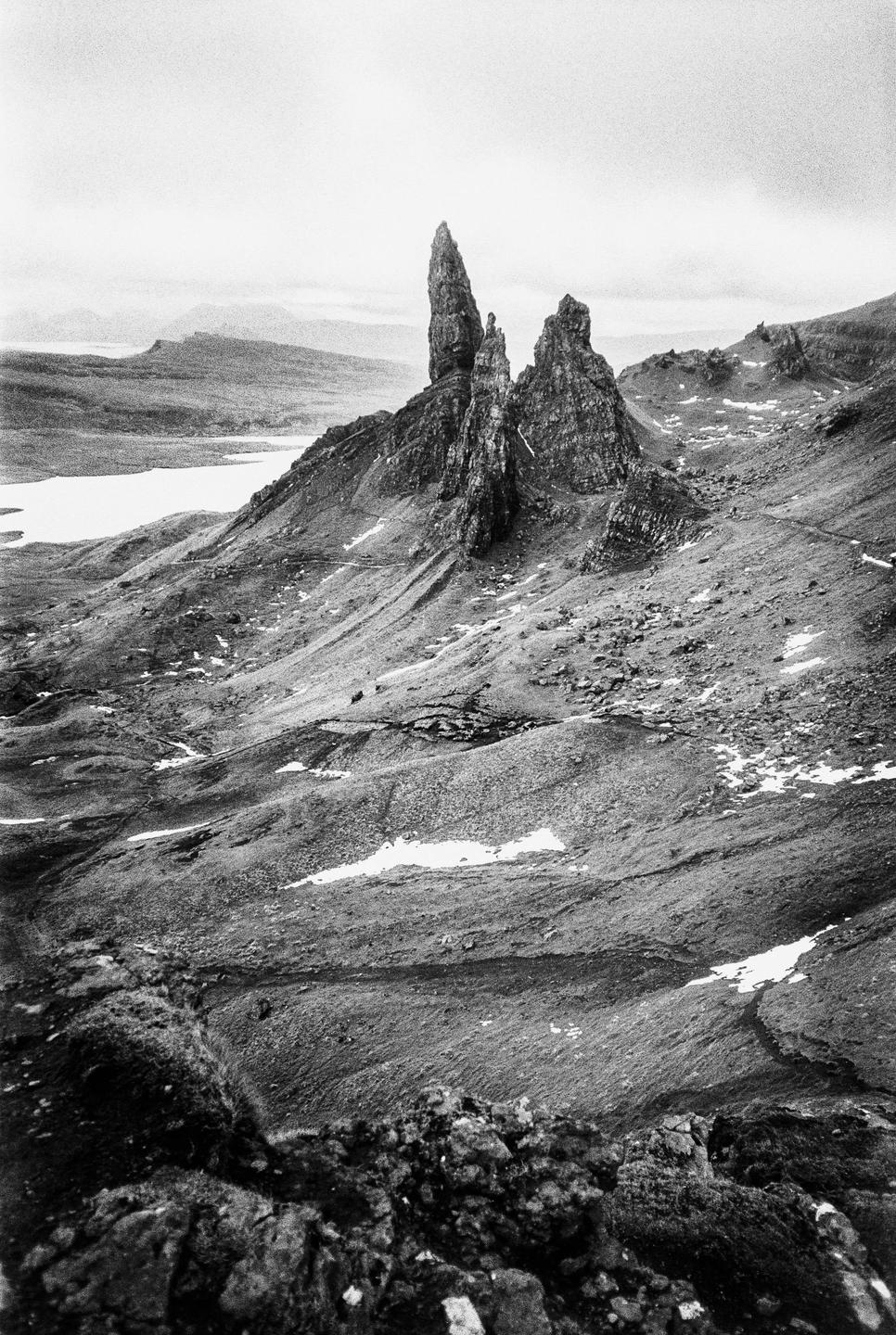 A black and white landscape shows a rocky mountain with a lake in the background under a cloudy sky.