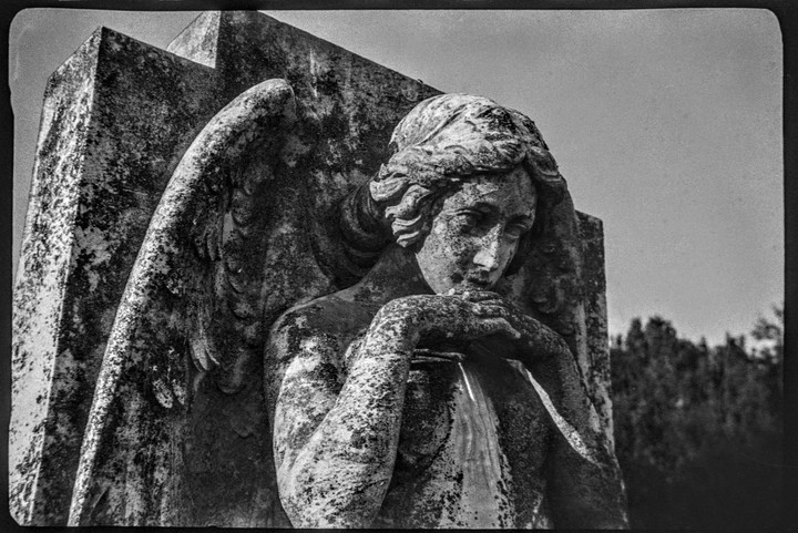 A weathered stone angel statue with wings is depicted in a close-up, looking downwards with hands clasped.