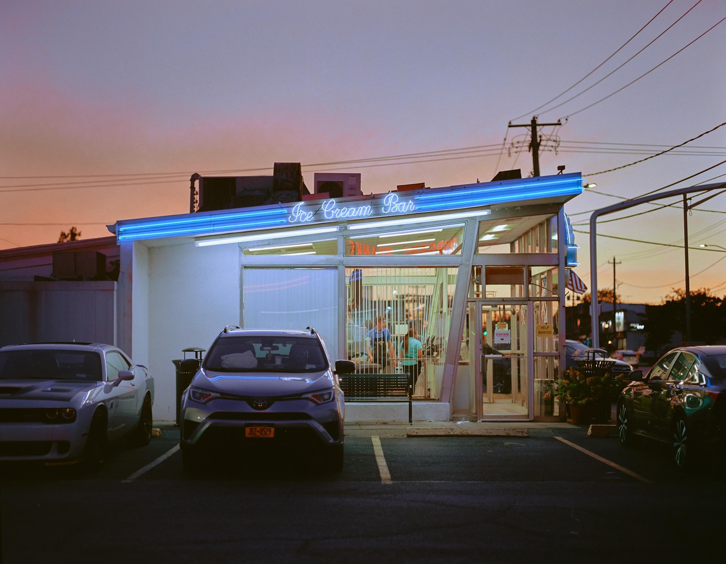 An ice cream bar with neon lights is visible at dusk, with cars parked in front.