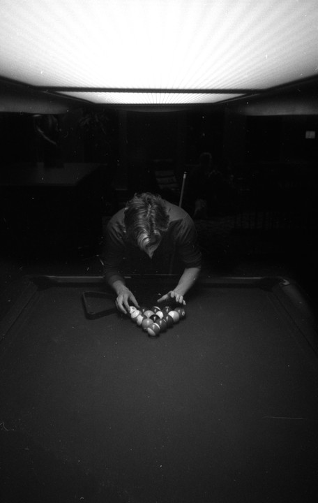 A man arranges billiard balls on a pool table under a bright overhead light.