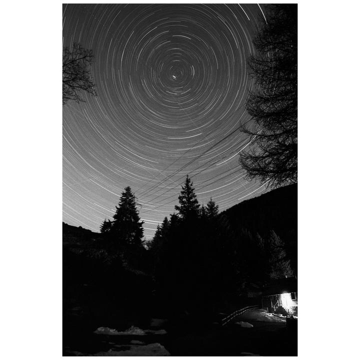 A long exposure photograph captures star trails circling Polaris above a dark forest and a lit cabin.