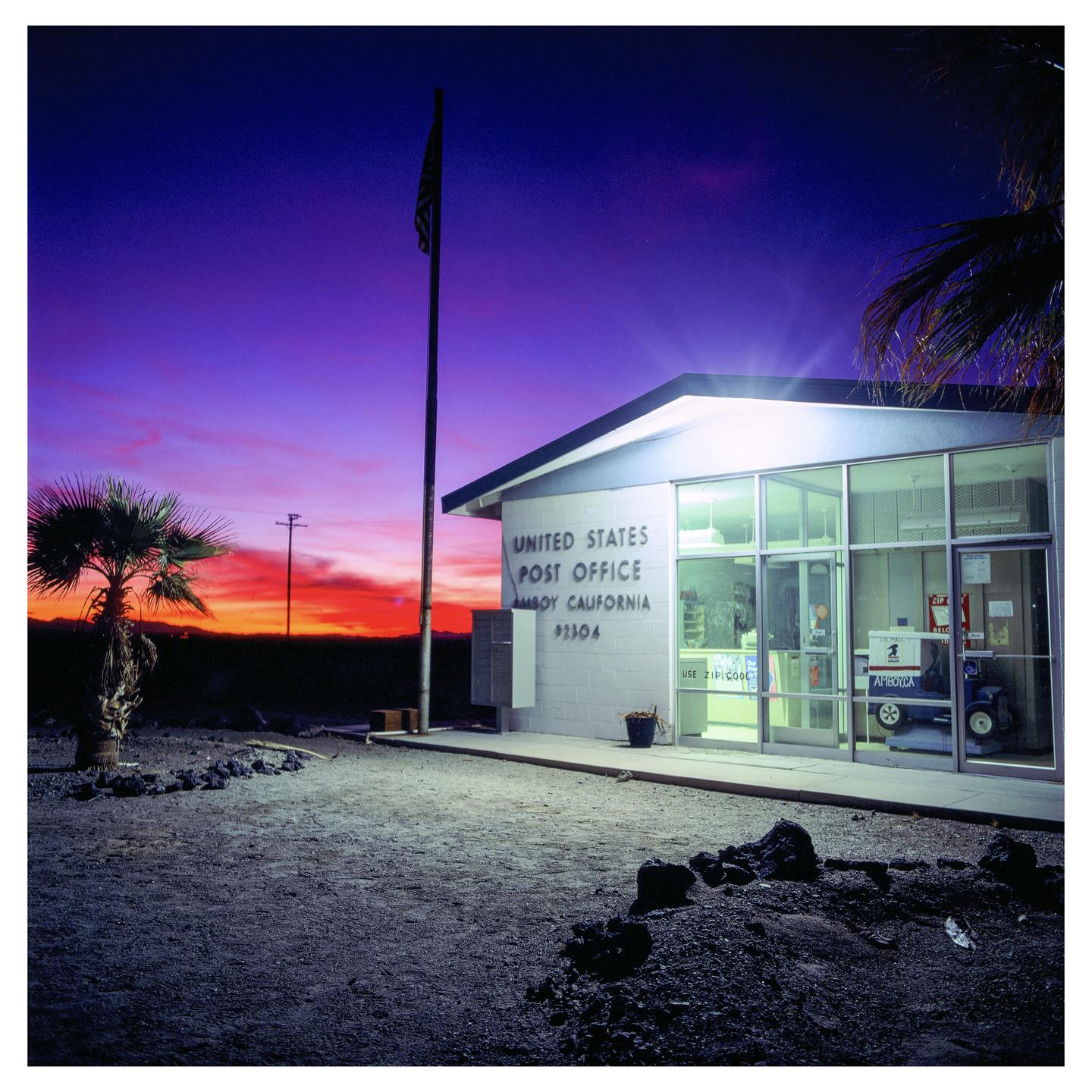 A United States Post Office building in Amboy, California is illuminated at dusk with a vibrant red and purple sky.
