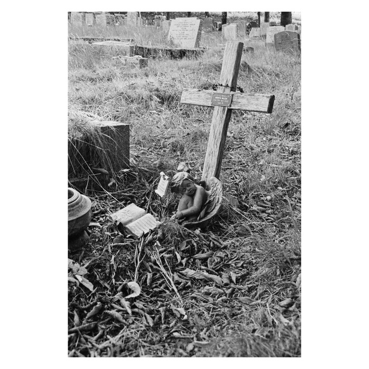A wooden cross and a cherub statue rest on a grave covered in dry leaves and grass.