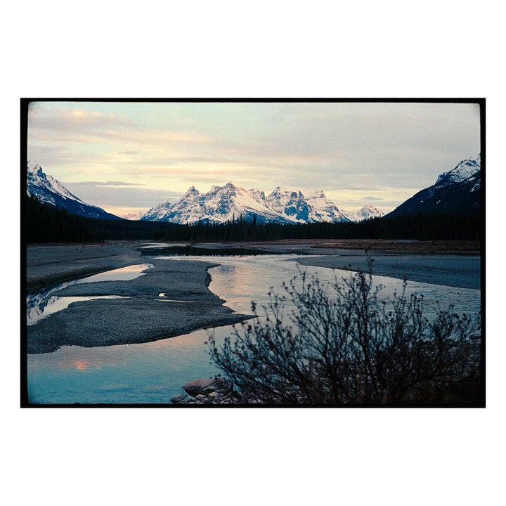 A river flows through a valley with snow-capped mountains and a forest under a cloudy sky.