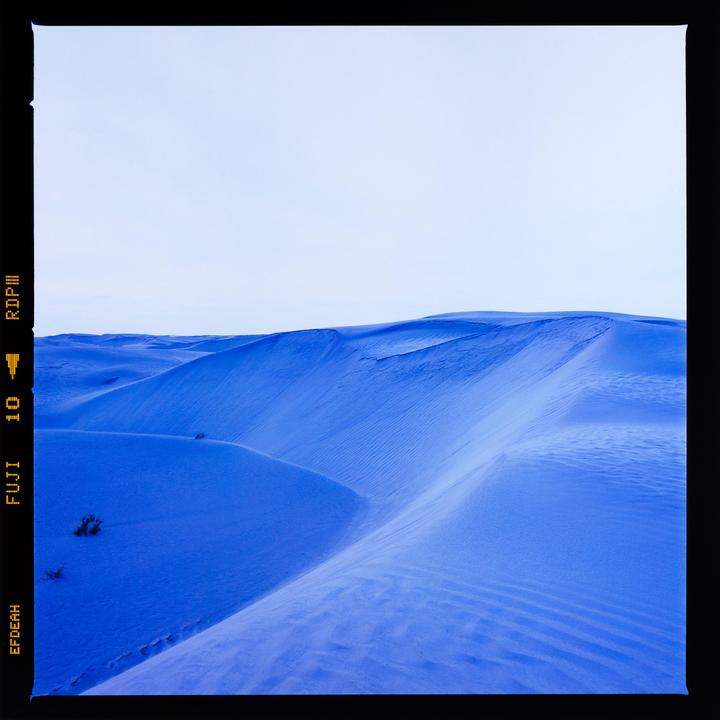 A vast expanse of blue sand dunes stretches under a light sky.