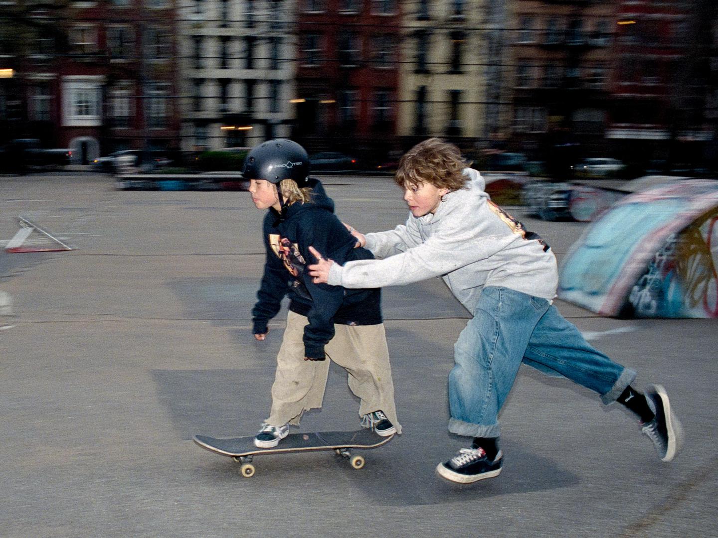 A boy in a helmet is on a skateboard while another boy pushes him from behind in a skatepark.