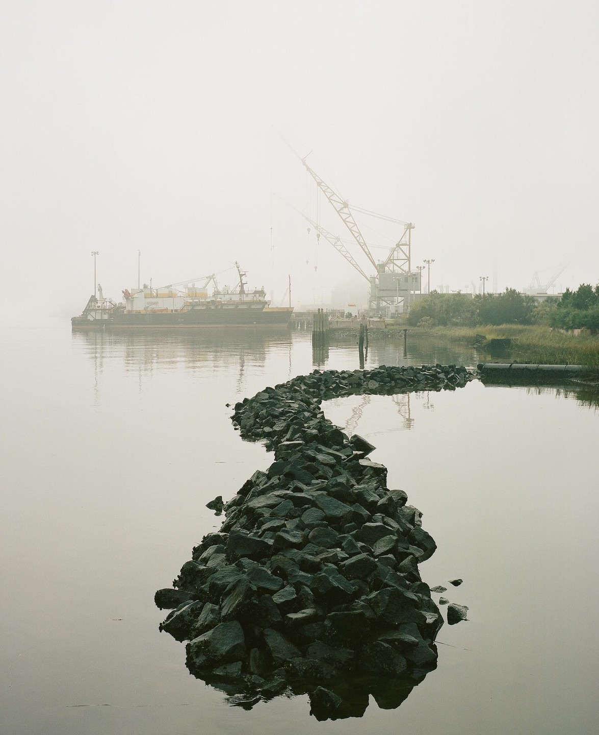 A rocky path leads into a foggy body of water with a ship and cranes in the background.