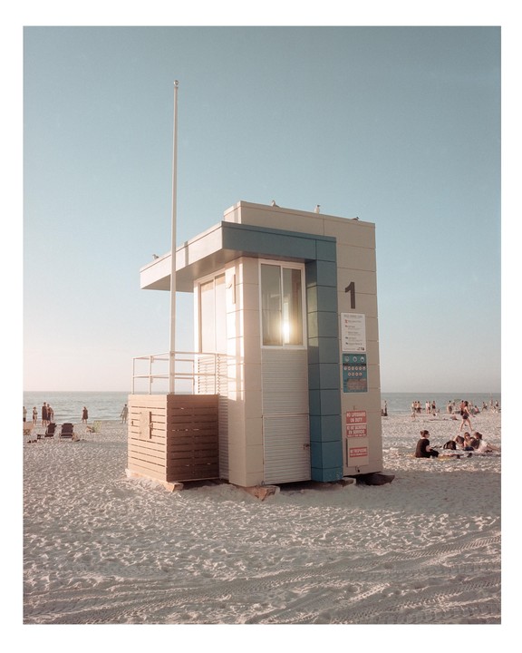 A lifeguard stand with the number one on its side stands on a sandy beach with people in the distance and the ocean under a clear sky.