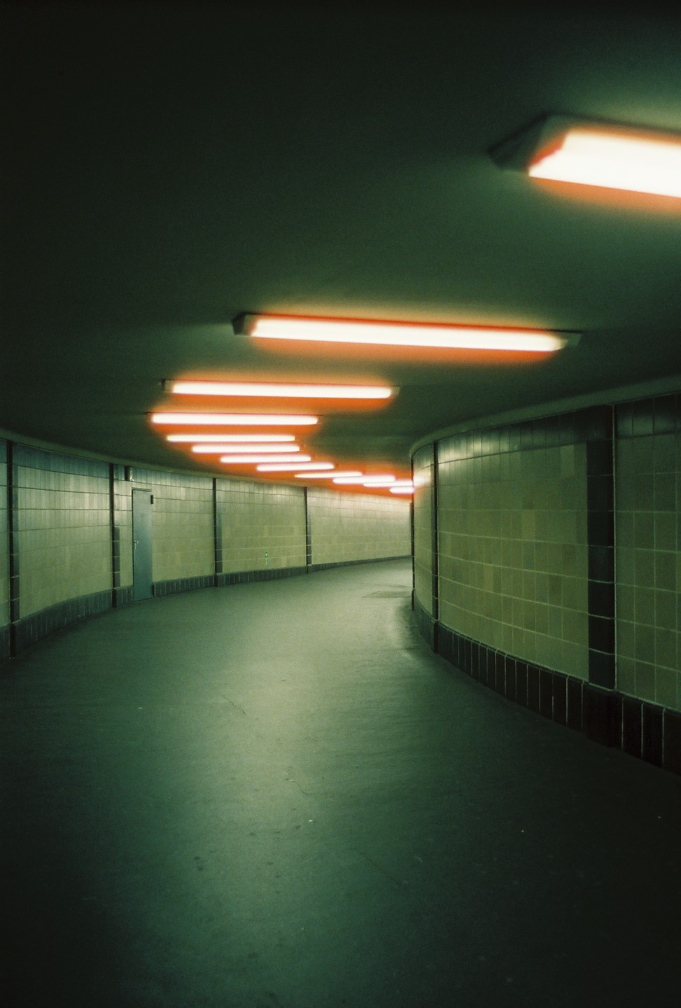 A long, curving hallway with tiled walls and a line of fluorescent lights on the ceiling.