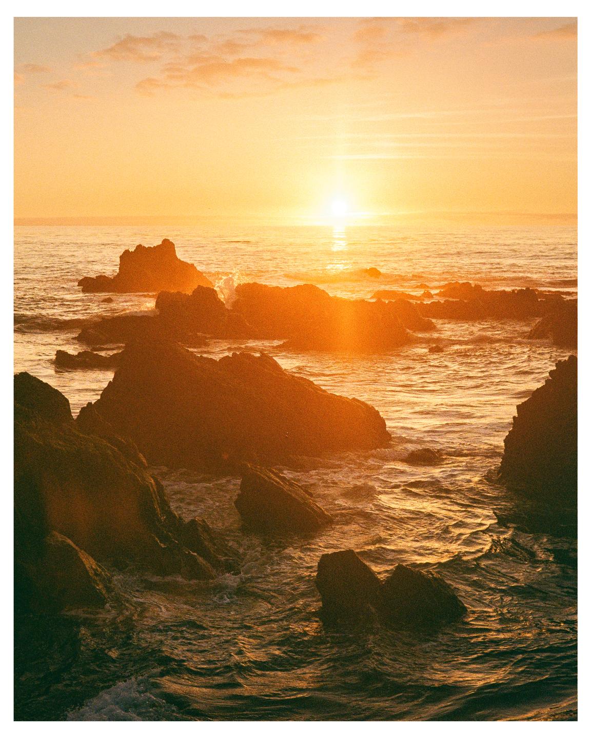 The sun sets over a rocky ocean with waves crashing against the shore.
