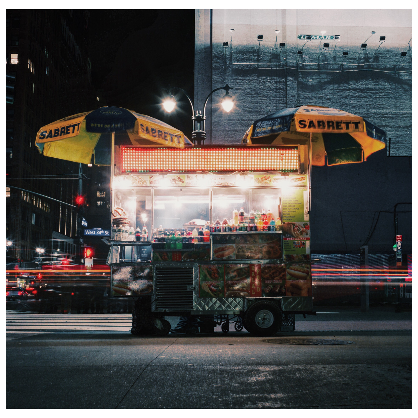 A brightly lit hot dog stand with two yellow umbrellas sits on a city street at night, with light trails from passing vehicles.