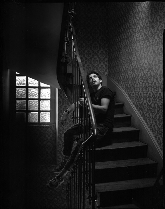A man sits on a wooden staircase with patterned wallpaper and looks up, next to a window with textured glass.