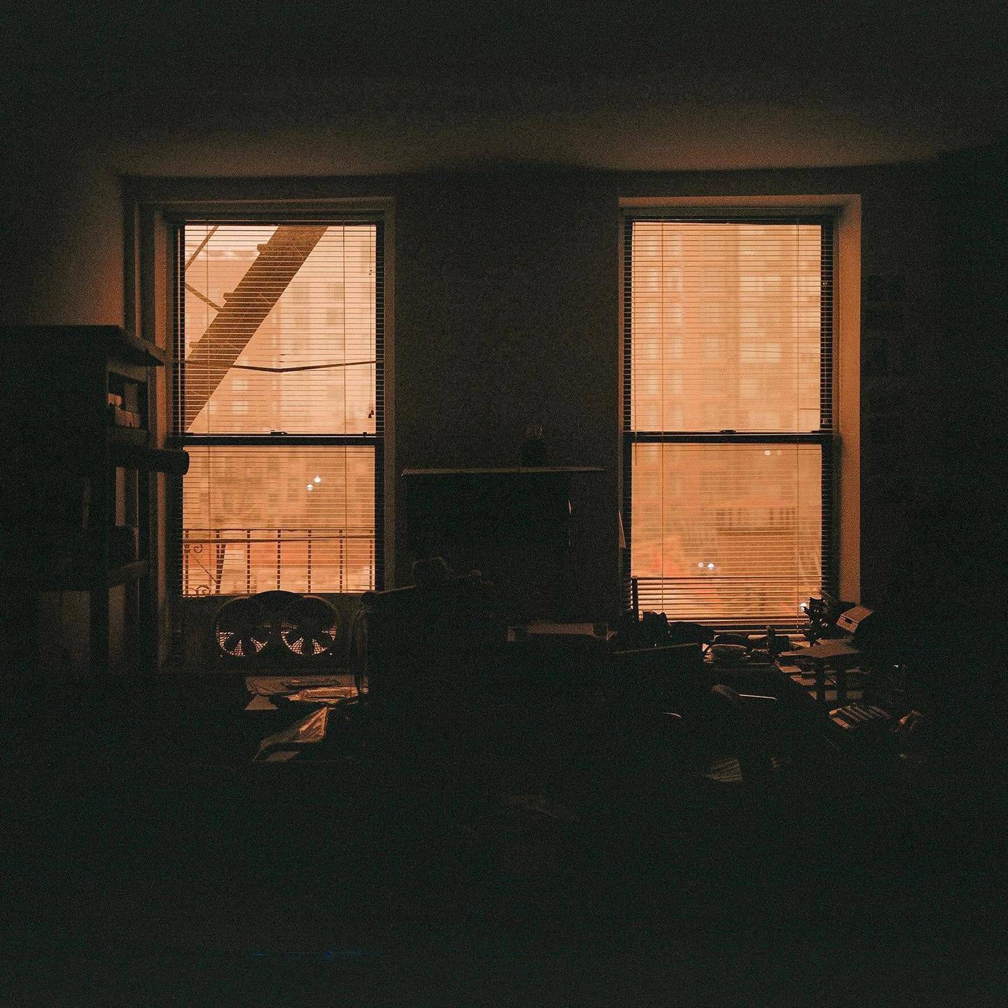A dark room is illuminated by orange light filtering through two windows with blinds.