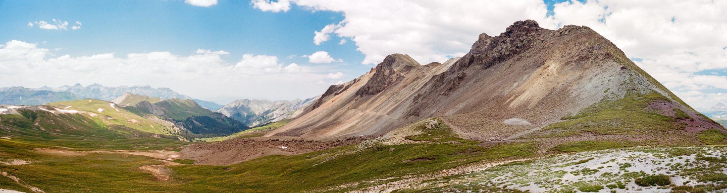 A panoramic landscape shows a mountain pass with green and brown slopes under a blue sky with white clouds.