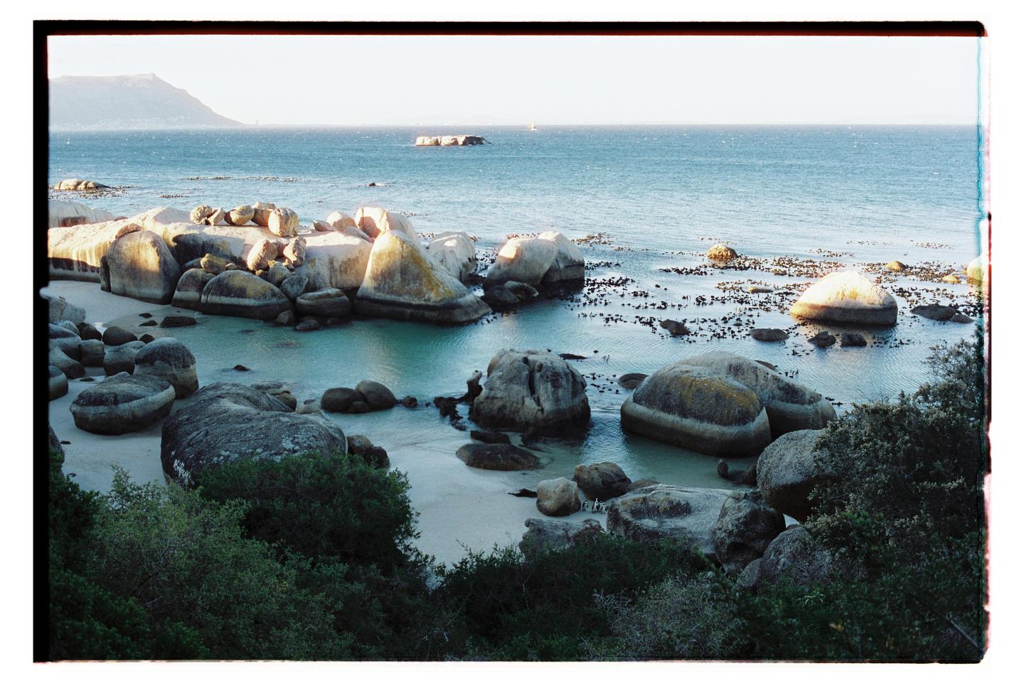 A beach with large boulders and calm water is visible, with a mountain in the distance.