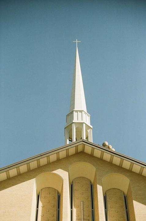 A white steeple with a cross on top rises above a brick building against a clear blue sky.
