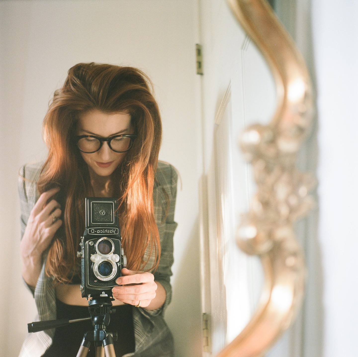 A woman with long red hair and glasses takes a self-portrait in a mirror with a camera on a tripod.