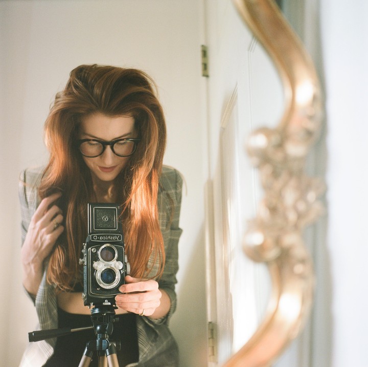 A woman with long red hair and glasses takes a self-portrait in a mirror with a camera on a tripod.