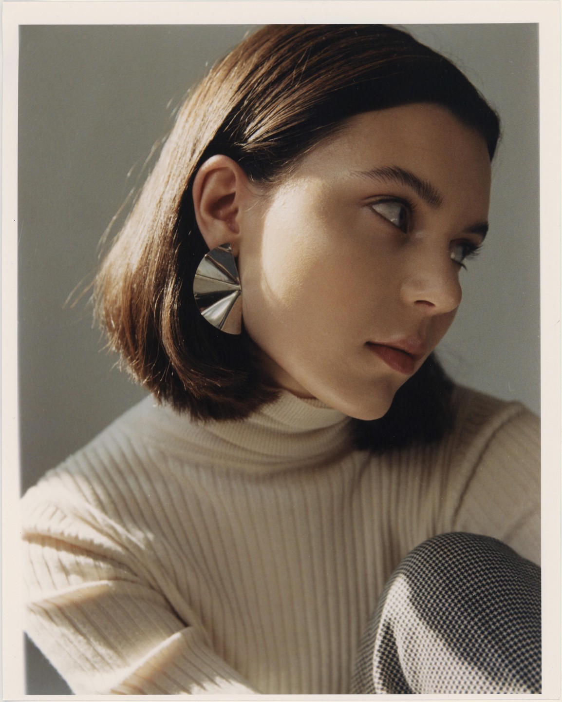 A woman with short brown hair and a cream turtleneck sweater looks to the right.