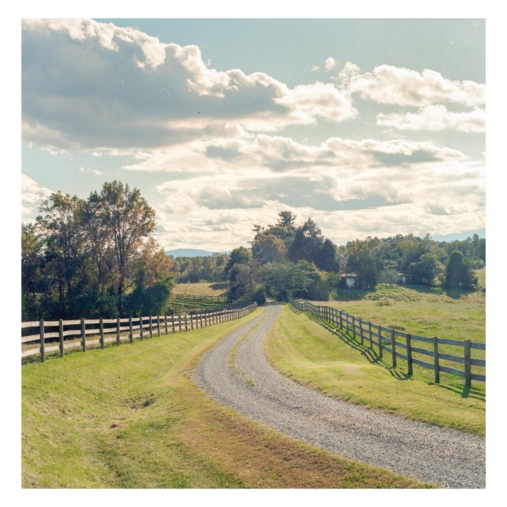 A gravel road curves through a green field with wooden fences on either side, leading towards distant trees and mountains under a cloudy sky.