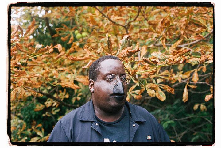A man wearing glasses and a bird-like nose and mouth stands in front of autumn foliage.