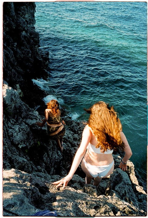 Two women climb down a rocky cliff towards the ocean.