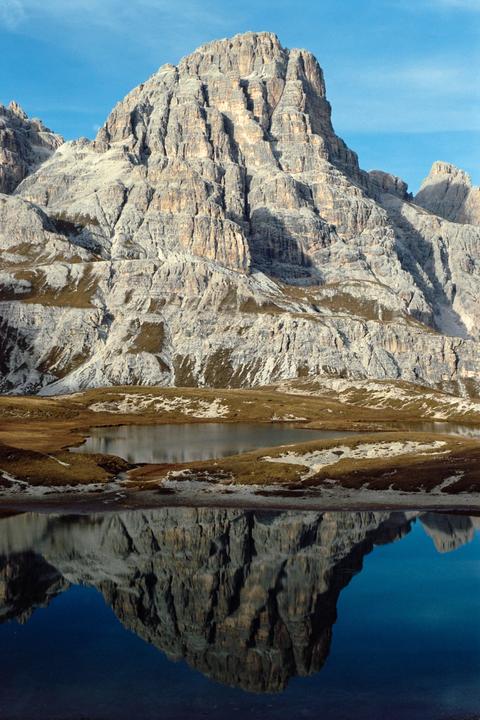 A large, rocky mountain is reflected in a still lake surrounded by grassy and snowy terrain.