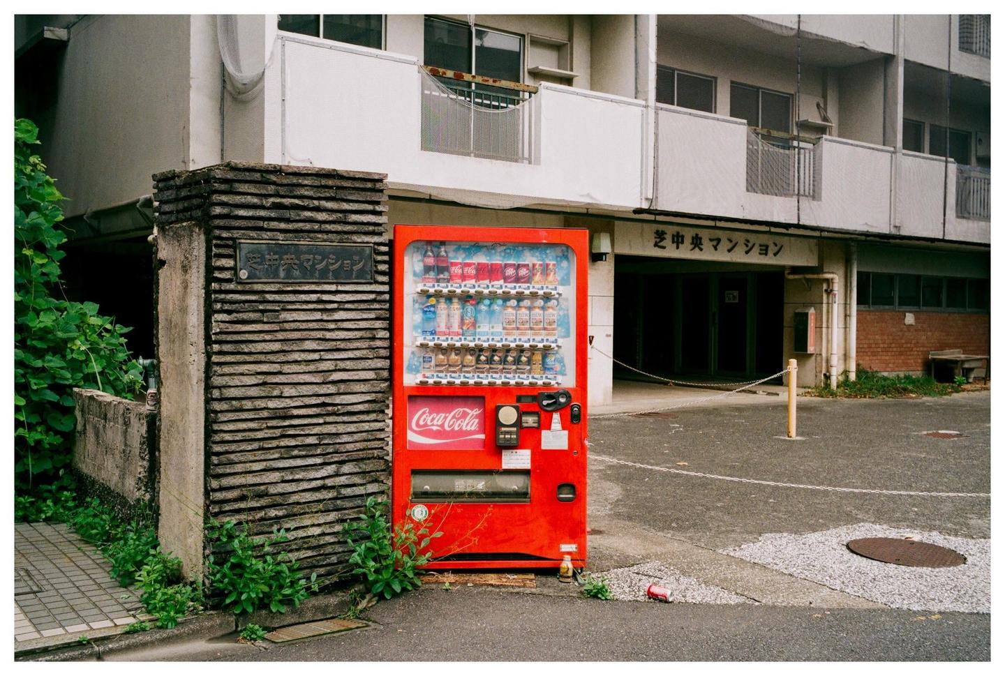 A red Coca-Cola vending machine stands next to a stone pillar with Japanese writing on it, in front of a building.
