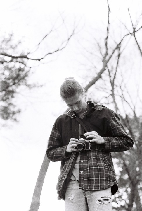 A man with a bun in his hair looks down at a camera he is holding in his hands.