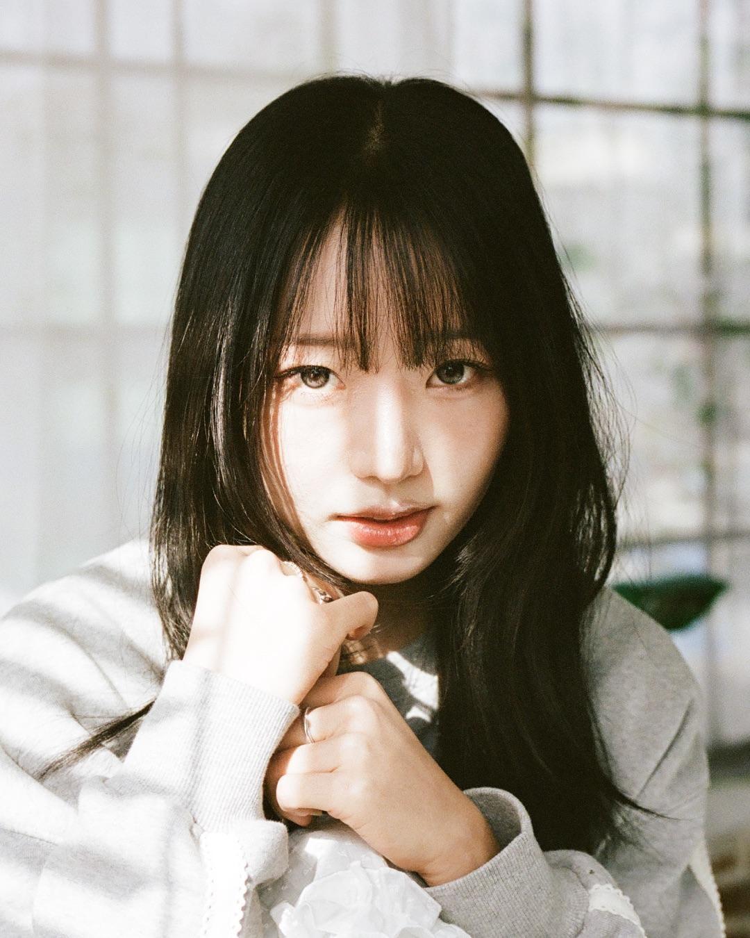 A woman with long dark hair and bangs looks directly at the viewer, with her hands clasped in front of her.