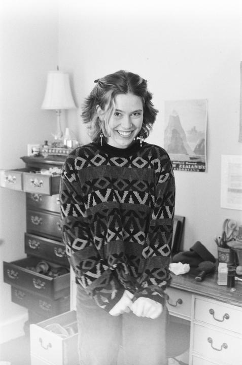 A smiling woman wearing a patterned sweater stands in a room with furniture and a poster on the wall.