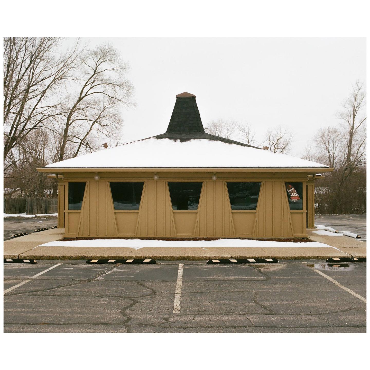 A hat-shaped building with a snow-covered roof stands in a parking lot with bare trees in the background.
