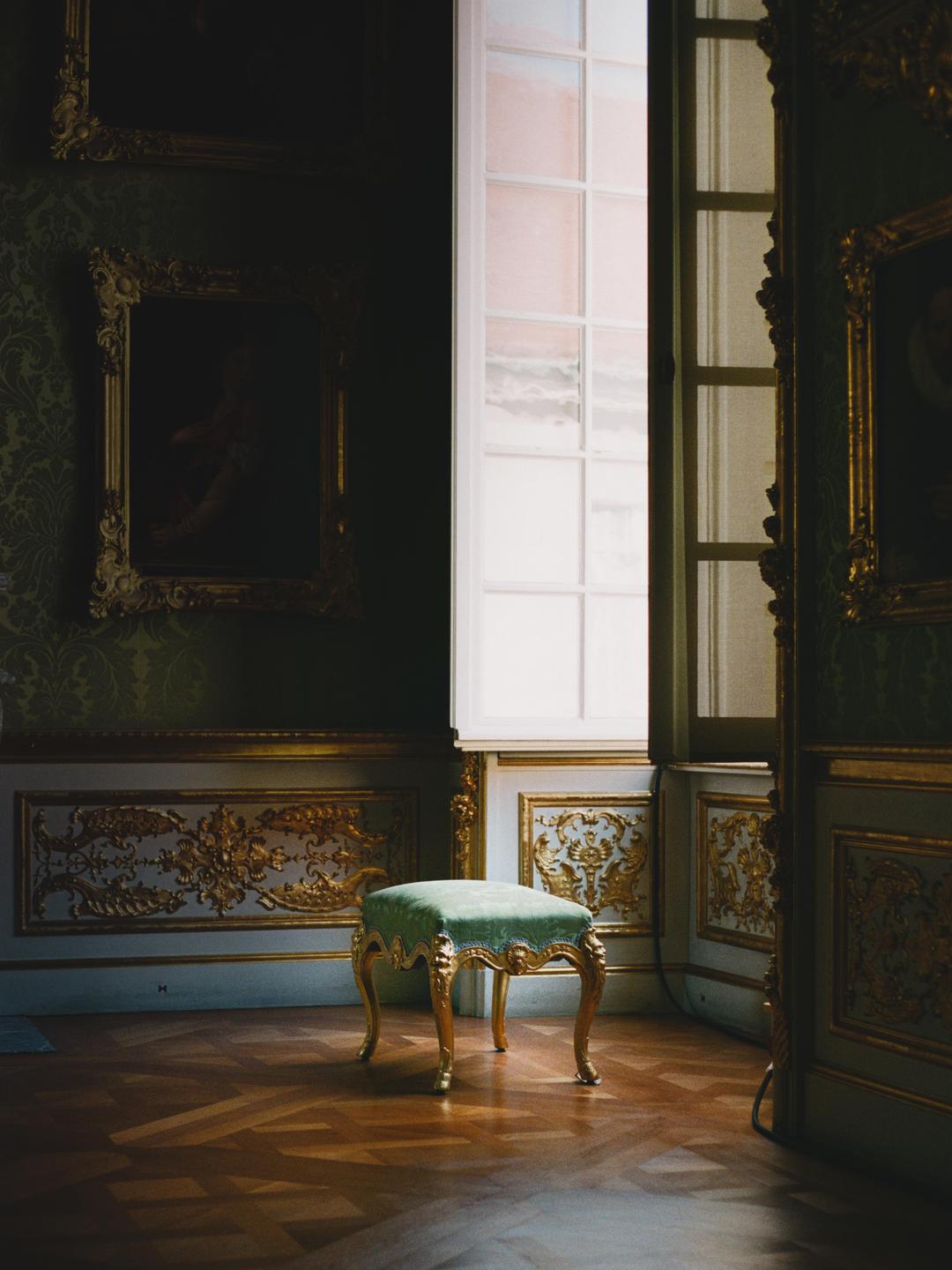 A green velvet stool with golden legs sits on a wooden floor in a room with ornate golden wall decorations and a large window.