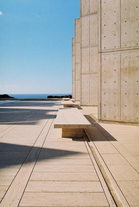 A concrete building with benches sits next to a paved area overlooking the ocean under a clear sky.
